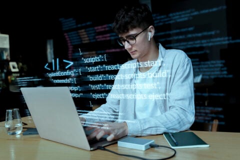 A person working on a laptop with code displayed on the screen, including commands for React scripts.