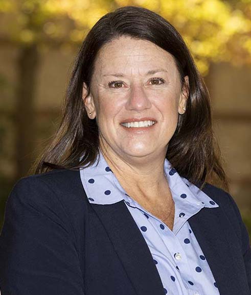 A woman wearing a navy blazer over a light blue polka dot blouse, standing outdoors with a blurred background.