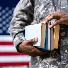 A soldier in camouflage holds a stack of books in front of an American flag.
