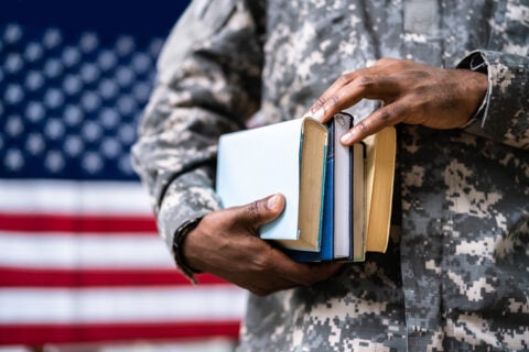 A soldier in camouflage holds a stack of books in front of an American flag.