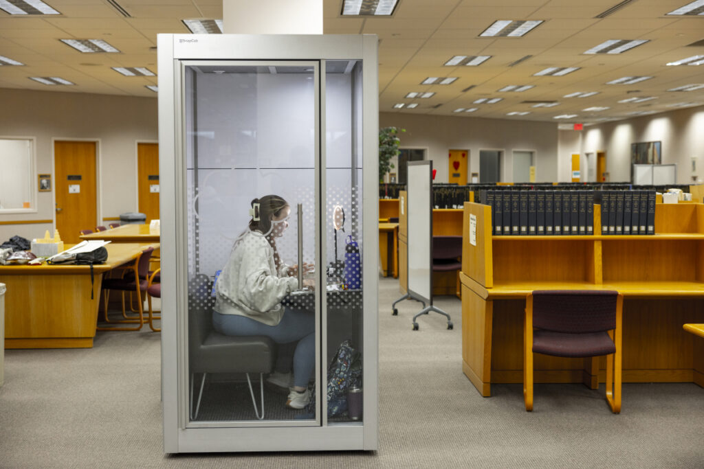 A person sitting inside a soundproof booth in a library, using a headset and working on a laptop.