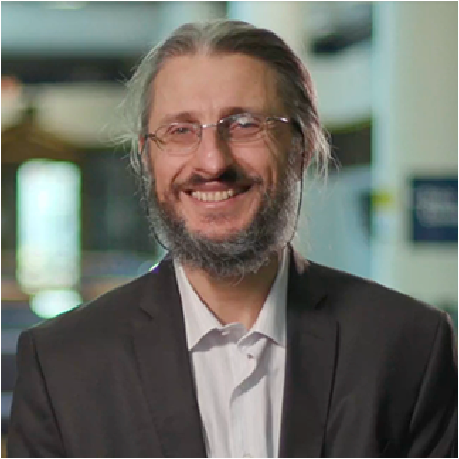 A person wearing a black suit and white shirt, standing in a well-lit indoor space.