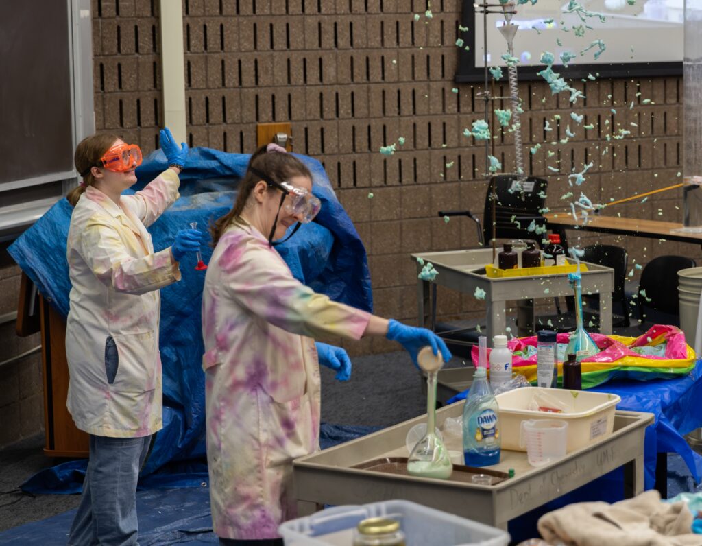 Two individuals in lab coats conduct a science experiment, causing a colorful foam to erupt from a flask in a classroom setting.