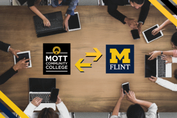 An overhead shot of eight people, mostly wearing business attire, sitting around a wooden conference table. Each person is focused on a laptop, tablet or smart phone. In the center are the Mott Community College logo and the University of Michigan-Flint logo. These logos are separated by two large yellow arrows pointing to MCC's logo and UM-Flint's logo, symbolizing a partnership or transfer agreement.