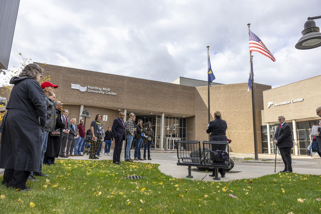 A group of people gathered outside the Harding Mott University Center, with flags flying and a cloudy sky above.
