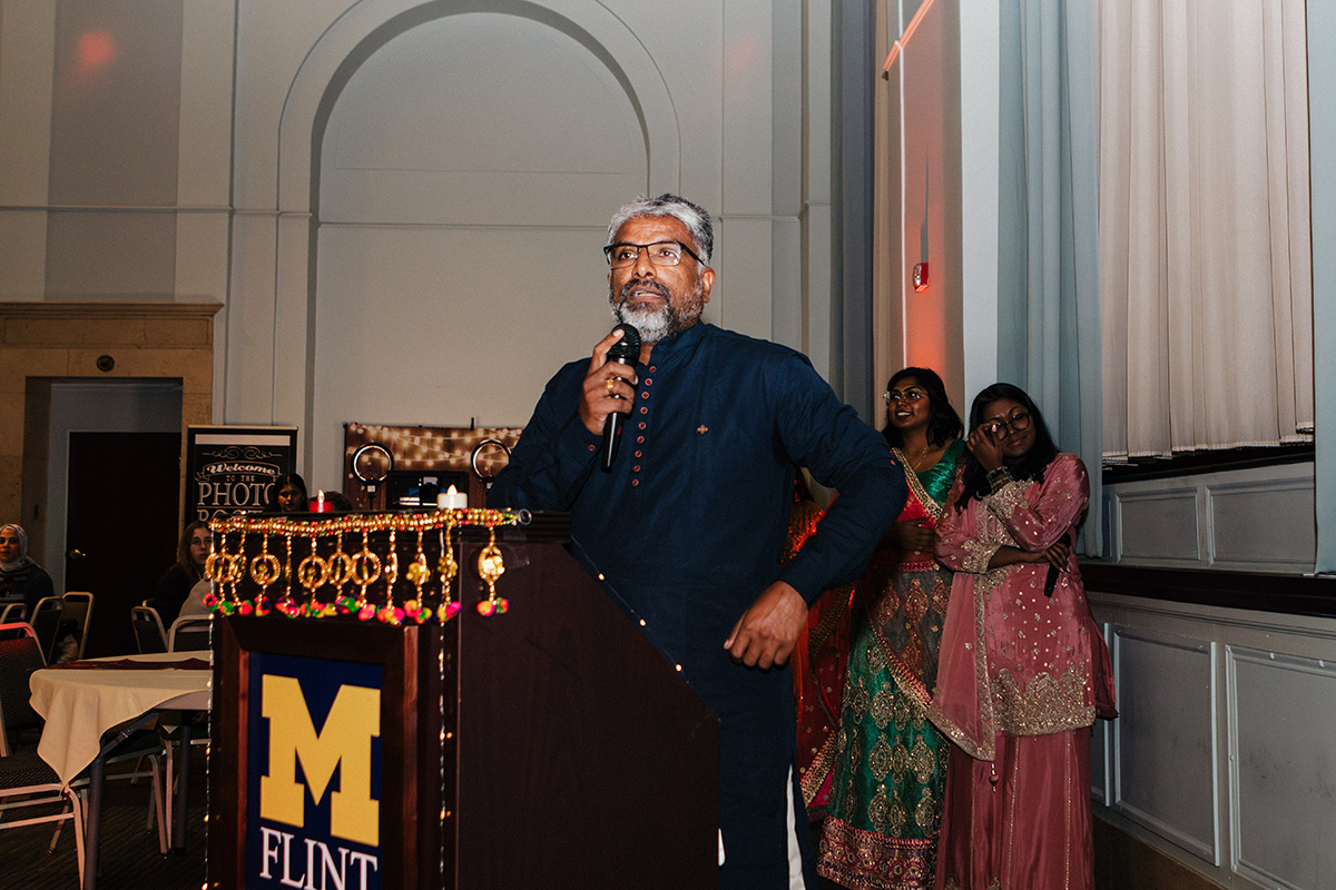 A person speaking at a podium decorated with a garland, with attendees in traditional attire in the background.