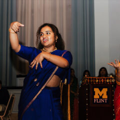 A woman in a blue sari performs a dance, while another woman in a red sari gestures nearby.