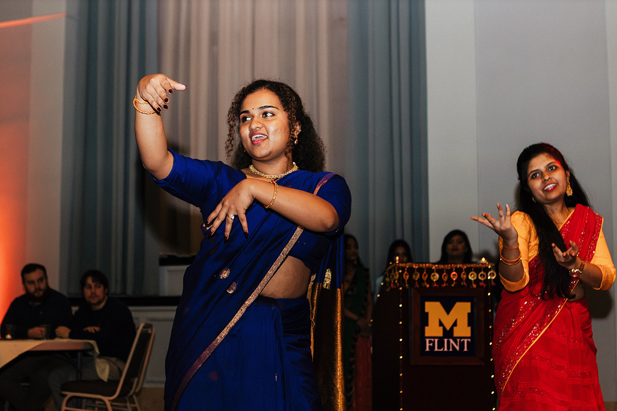 A woman in a blue sari performs a dance, while another woman in a red sari gestures nearby.