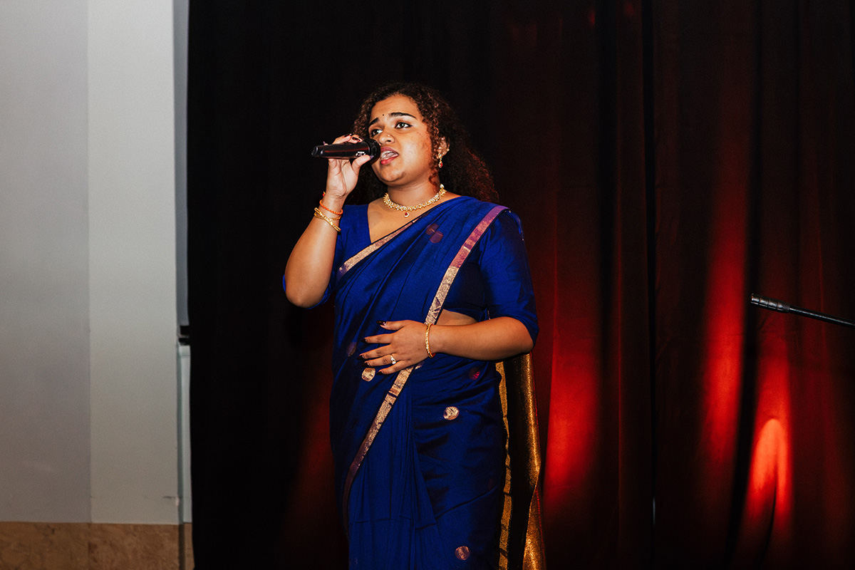 A woman in a blue sari sings into a microphone on stage, with a dark curtain and red lighting in the background.