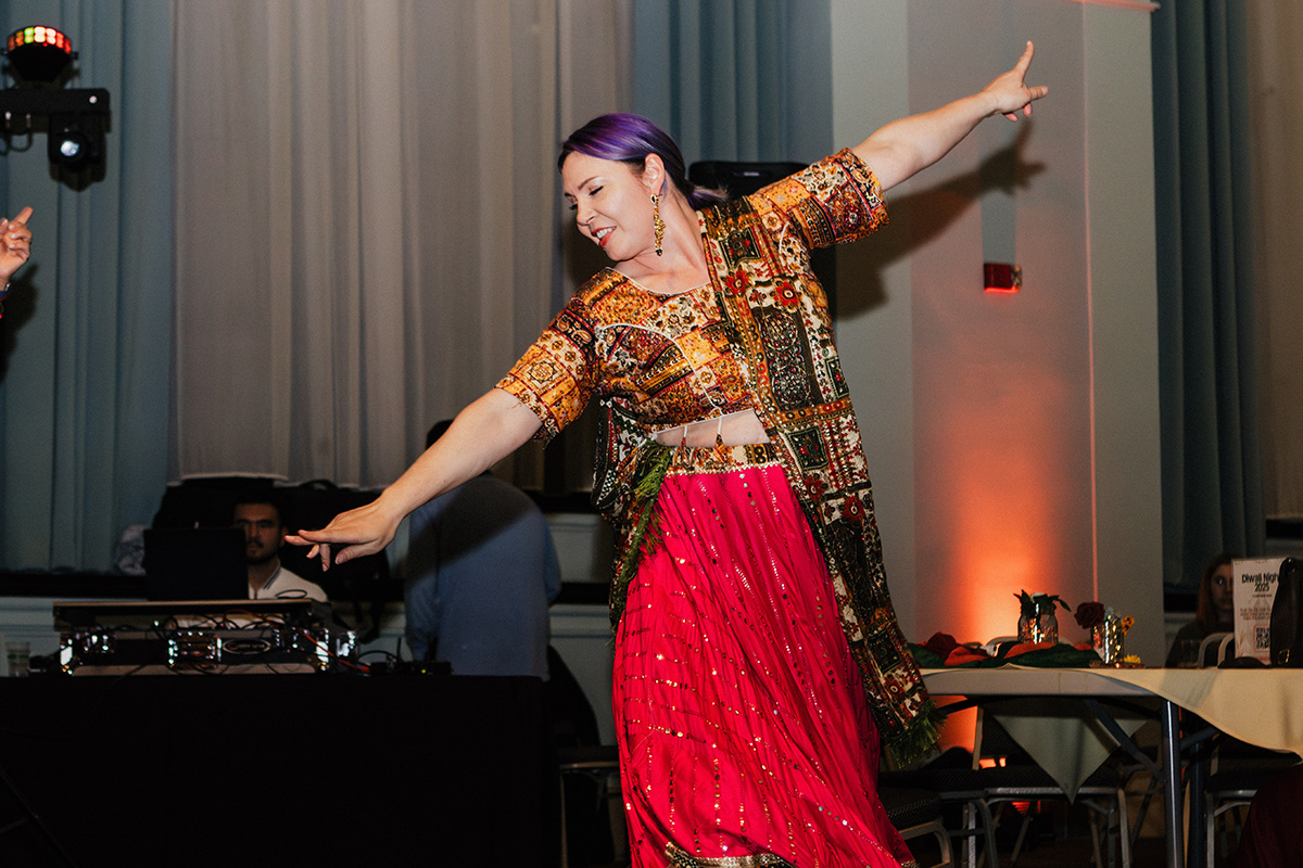A person in a colorful traditional outfit dances joyfully, with one arm raised and a vibrant red skirt flowing.