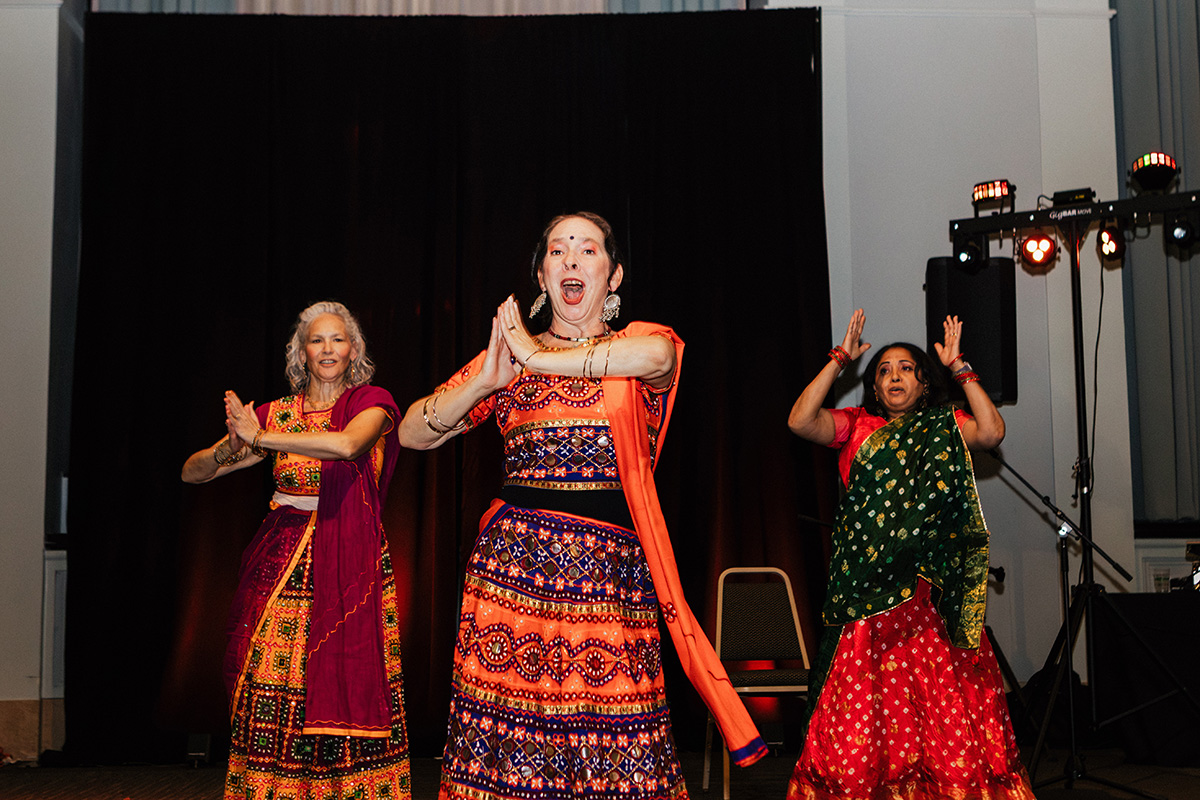 Three women in colorful traditional outfits are performing a dance on stage, with a dark backdrop and stage lights.
