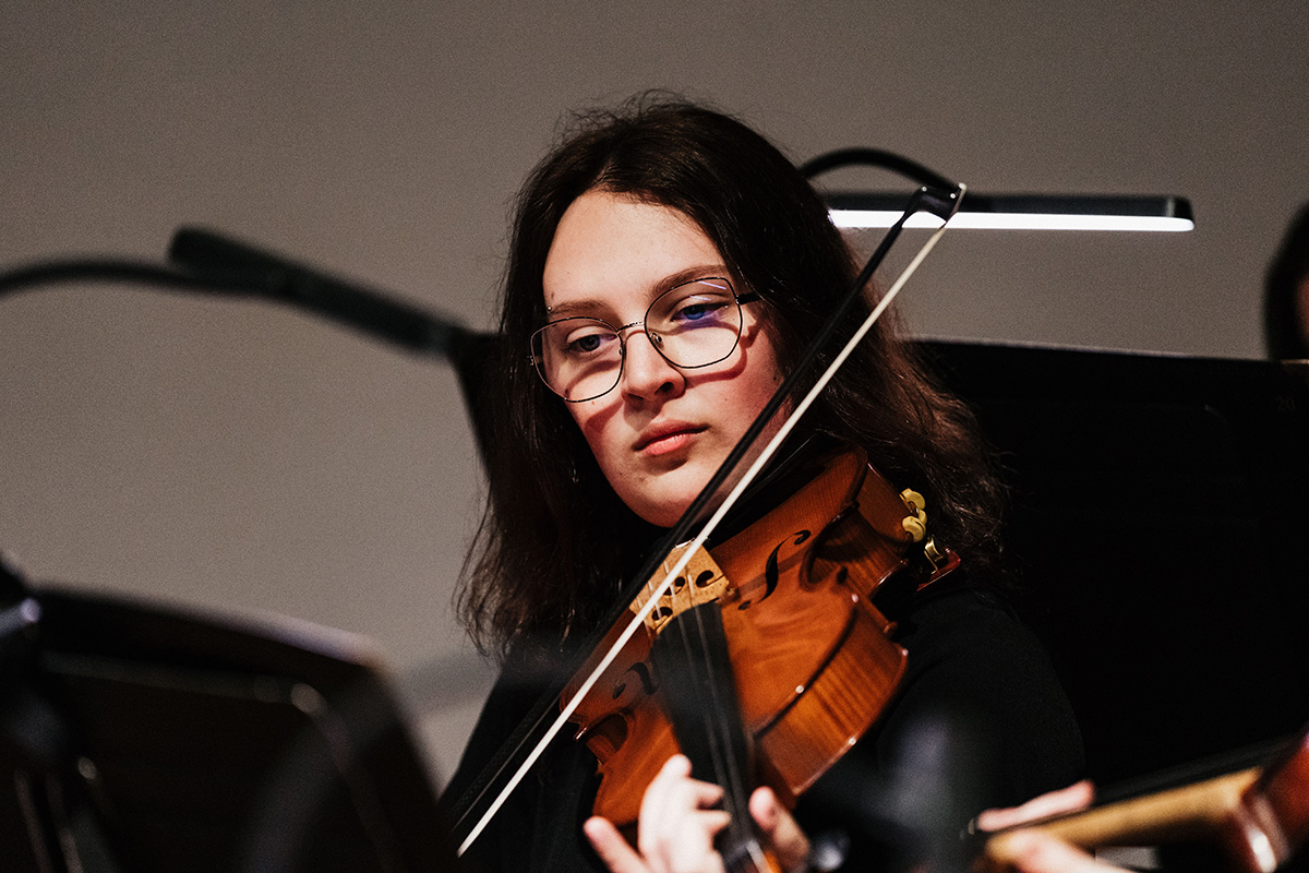 A musician playing a violin under a focused light, with music stands visible in the background.