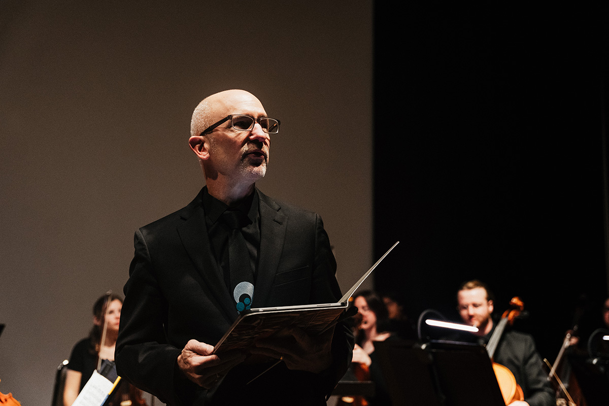 A conductor in a black suit holds a music score while leading an orchestra in the background.