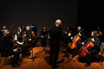 A conductor leads an orchestra of musicians playing string instruments in a dimly lit performance space.