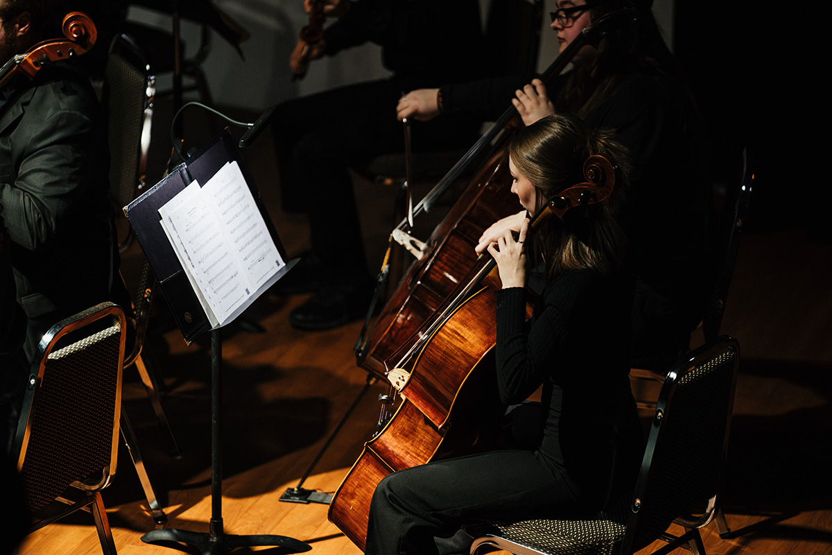 A musician playing a cello in an orchestra setting, with sheet music on a stand nearby.