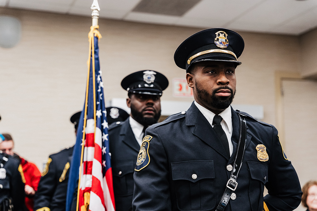 FLINT_Veterans Day 2025_0015 A police officer in uniform stands in front of an American flag, with other officers visible in the background.