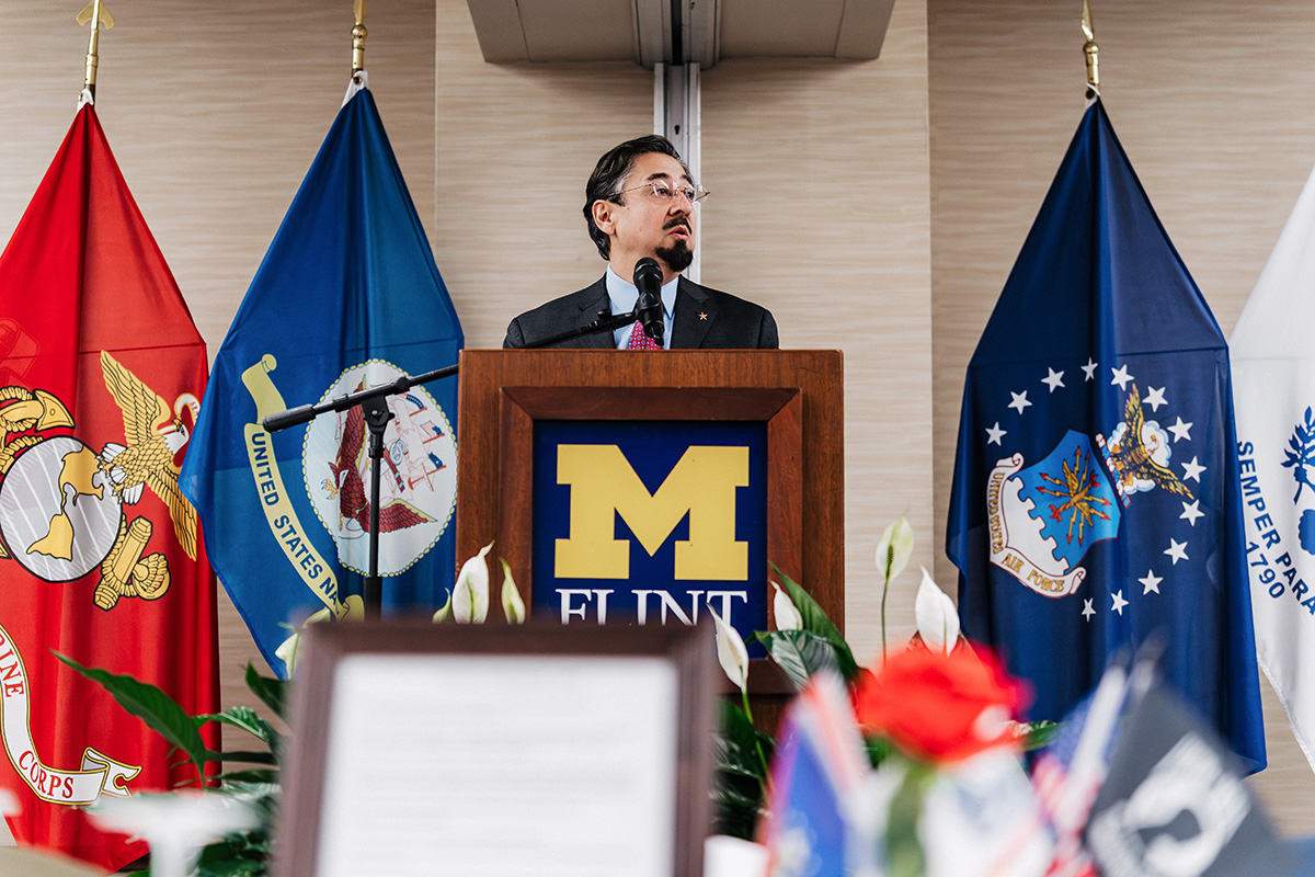 FLINT_Veterans Day 2025_0037 A speaker stands behind a podium with the "M FLINT" logo, flanked by flags representing the Marine Corps, Navy, and Air Force.