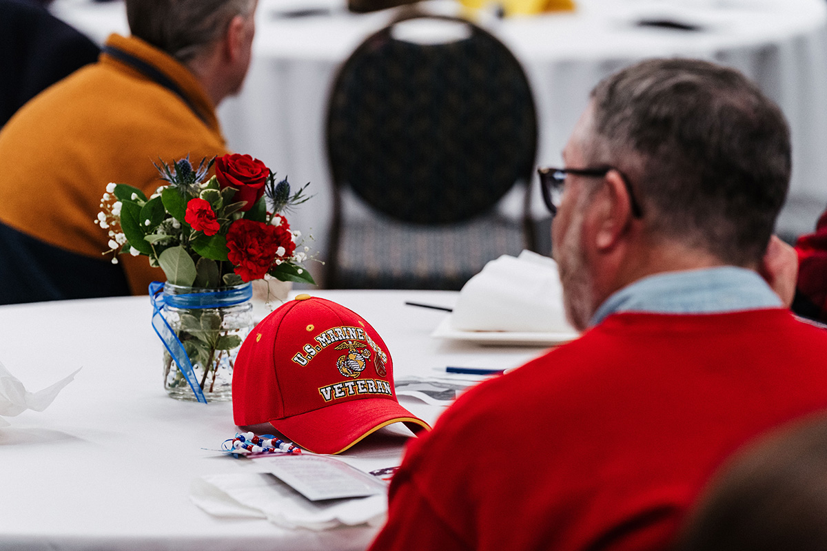 FLINT_Veterans Day 2025_0044 A red U.S. Marine Corps veteran cap rests on a table next to a floral arrangement and other items.
