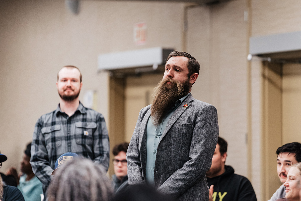 FLINT_Veterans Day 2025_0052 A man with a long beard stands in a meeting room, wearing a gray blazer and a blue shirt, while others are seated around him.