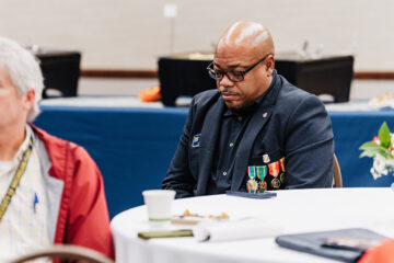 A man wearing a dark blazer with medals sits at a table with a coffee cup and papers, surrounded by other attendees.