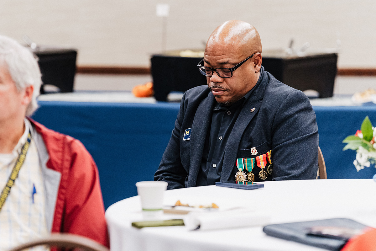 A man wearing a dark blazer with medals sits at a table with a coffee cup and papers, surrounded by other attendees.