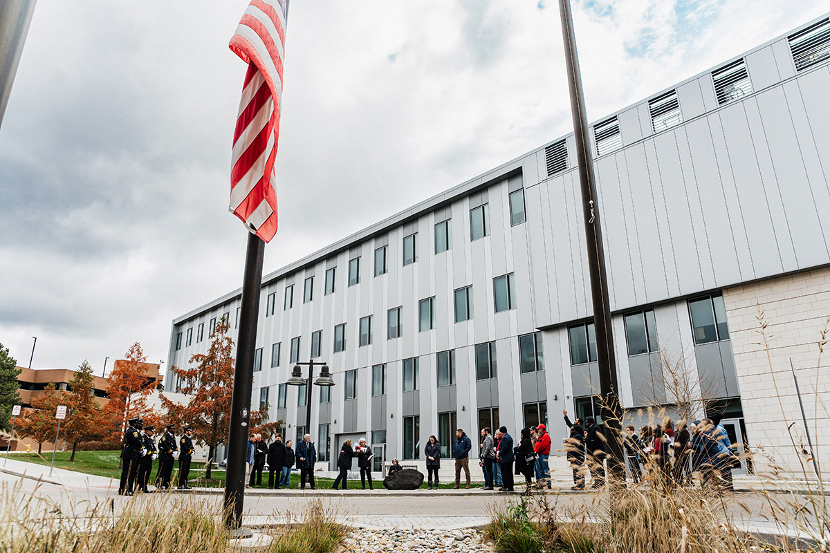 FLINT_Veterans Day 2025_0080 A group of people gathered outside a modern building, with a flagpole displaying an American flag in the foreground.