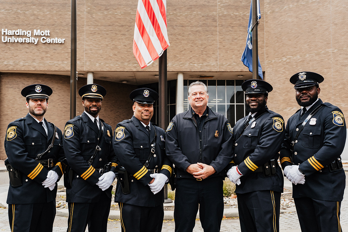 FLINT_Veterans Day 2025_0087 A group of six police officers in uniform standing in front of the Harding Mott University Center, with an American flag in the background.