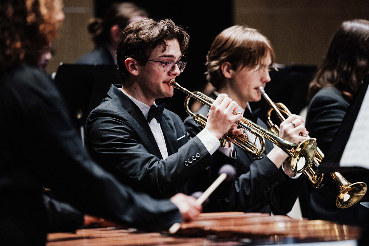 A group of musicians in formal attire playing trumpets in an orchestra setting.