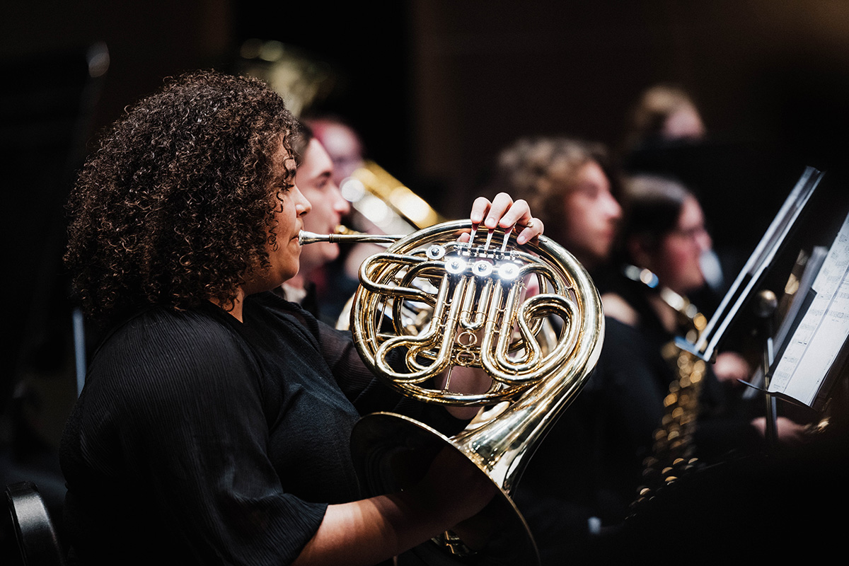 A musician playing a French horn in an orchestra setting, with other musicians blurred in the background.