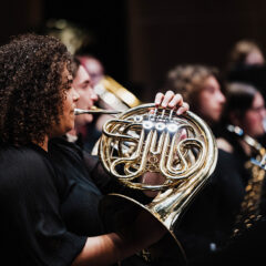 A musician playing a French horn in an orchestra setting, with other musicians visible in the background.