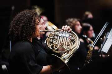 A musician playing a French horn in an orchestra setting, with other musicians visible in the background.