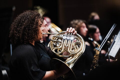 A musician playing a French horn in an orchestra setting, with other musicians visible in the background.