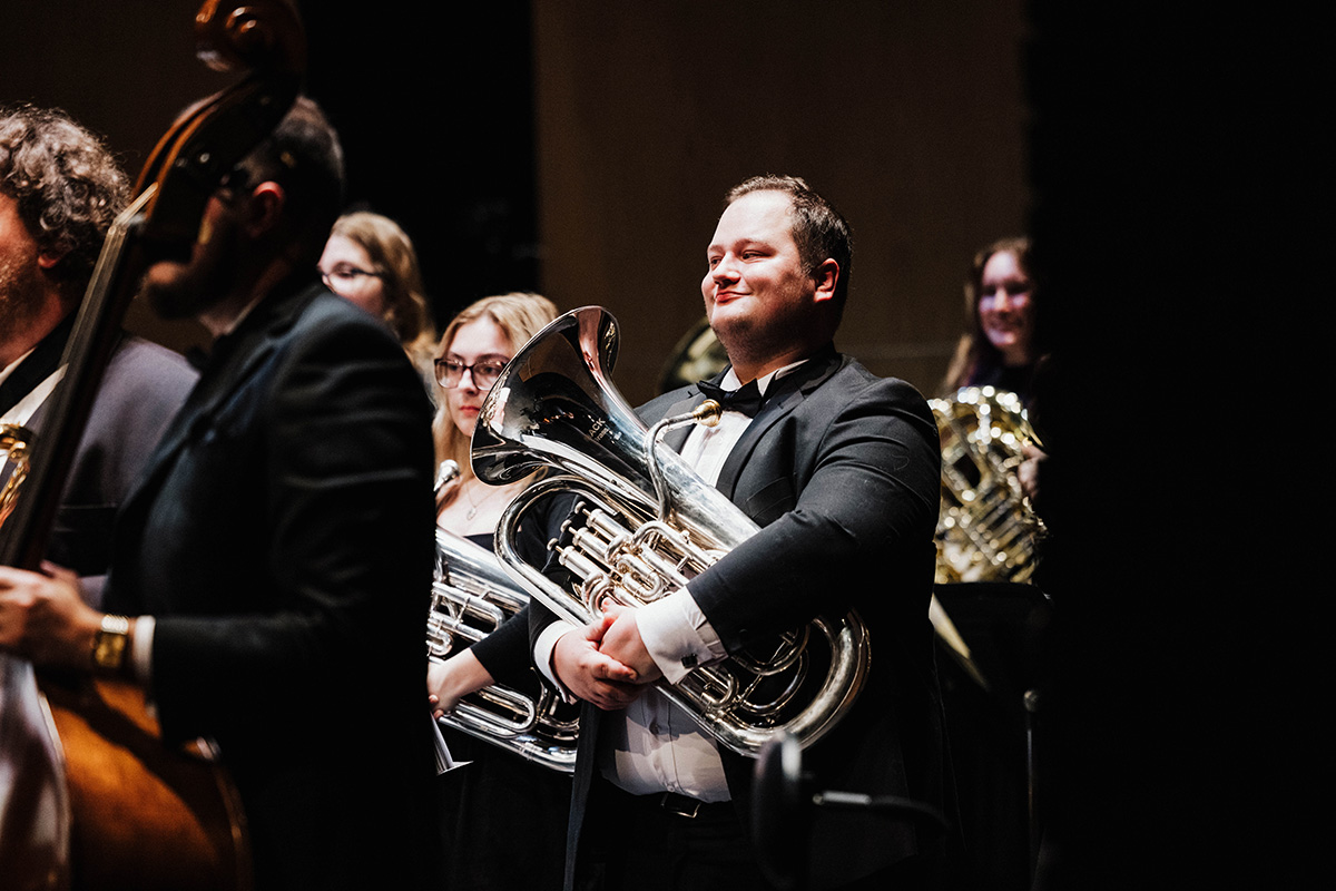 A musician in formal attire holds a tuba while standing among other musicians in an orchestra setting.