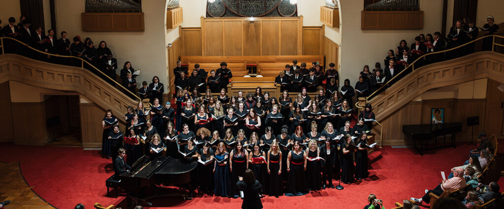 A large choir of singers in formal attire performs on stage, accompanied by a pianist, in a concert hall setting.