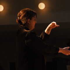 A person in a black outfit is conducting, with their right hand raised and fingers poised, under soft lighting.