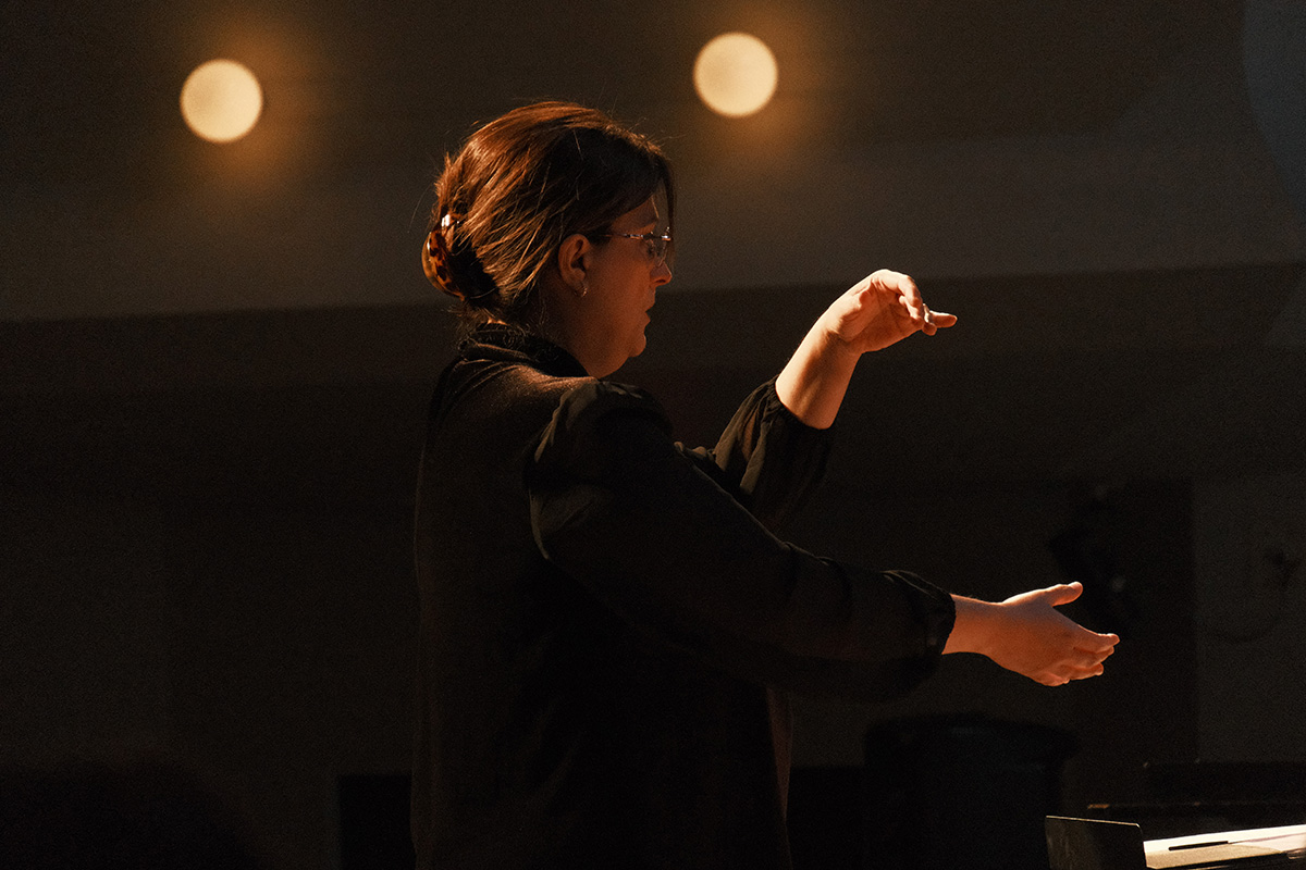 A person in a black outfit is conducting, with their right hand raised and fingers poised, under soft lighting.