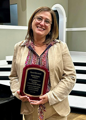 A person holding a plaque with a dark wooden frame and gold text, standing in a formal setting.