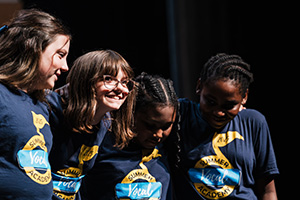 A group of children wearing matching blue t-shirts with a yellow logo, standing close together on stage.
