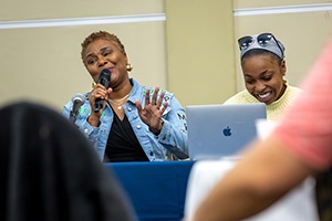 A person speaking into a microphone while gesturing with their hand, seated at a table with a laptop.