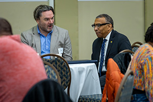 Two people seated at a table in a conference setting, engaged in conversation.