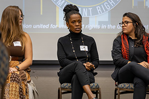 Three women seated on chairs during a panel discussion, with a presentation screen in the background.