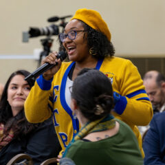 A person in a yellow and blue cardigan is speaking into a microphone at a gathering, with an audience in the background.