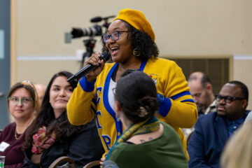 A person in a yellow and blue cardigan is speaking into a microphone at a gathering, with an audience in the background.