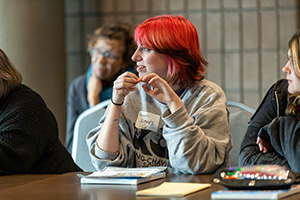 A person with red hair sits at a table, holding a pen and looking thoughtful, surrounded by others in a meeting setting.