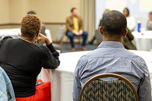 A group of people seated at tables in a meeting room, with two individuals in the foreground facing away from the camera.