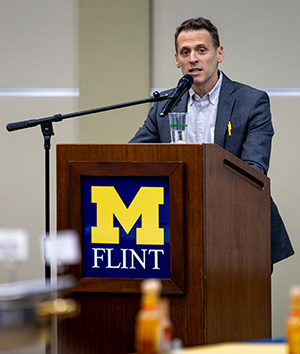 A person speaking at a podium with a sign that reads "M FLINT" and a microphone.