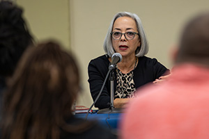 A woman seated at a table with a microphone, speaking to an audience.