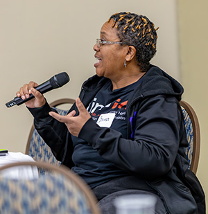 A person wearing a black hoodie is speaking into a microphone while gesturing with their hands.