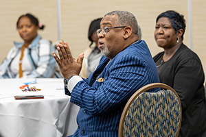 A man in a blue pinstripe suit sits at a table, hands clasped, with other attendees in the background.