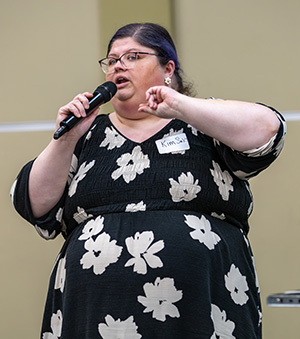 A person wearing a black floral dress holds a microphone and speaks at an event. A name tag is visible on their clothing.